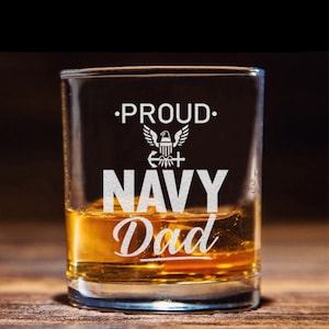 May include: A clear glass shot glass with the words "Proud Navy Dad" etched on the front. The glass is filled with amber liquid and has a white eagle emblem with an anchor and cross.