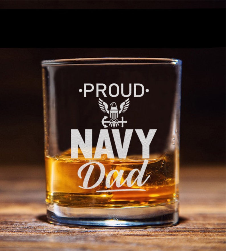 May include: A clear glass shot glass with the words "Proud Navy Dad" etched on the front. The glass is filled with amber liquid and has a white eagle emblem with an anchor and cross.