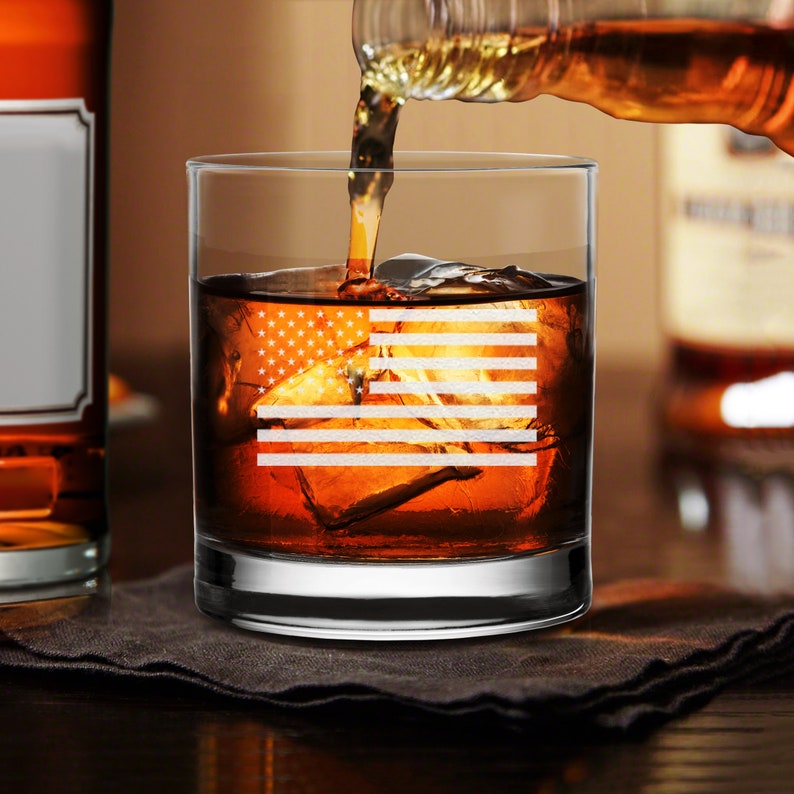 Puede incluir: Un vaso de rocas de vidrio transparente con un dise&ntilde;o de bandera estadounidense grabado. El vaso est&aacute; lleno de l&iacute;quido &aacute;mbar y cubitos de hielo.