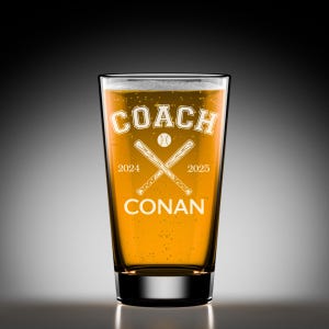 May include: A pint glass filled with a golden liquid, possibly beer, featuring the word "COACH" above crossed baseball bats and a baseball. The text "2024" and "2025" flank the bats, with "CONAN" below. The glass has a black base.