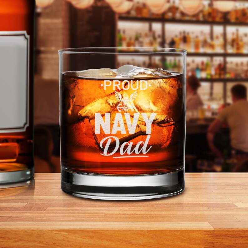 May include: A clear glass tumbler with a design that says "Proud Navy Dad" with an eagle graphic. The glass is filled with amber liquid and ice cubes.
