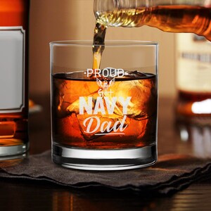 May include: A clear glass with the words "Proud Navy Dad" etched on the side. The glass is filled with amber liquid and ice cubes.