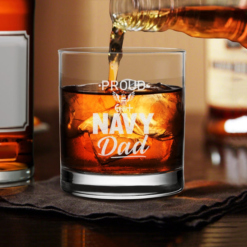 May include: A clear glass with the words "Proud Navy Dad" etched on the side. The glass is filled with amber liquid and ice cubes.