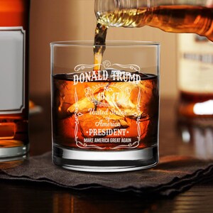 Puede incluir: Un vaso de rocas de vidrio transparente con el texto "DONALD TRUMP No. 45 & 47 United States of America &bull;PRESIDENT&bull; MAKE AMERICA GREAT AGAIN" grabado en el lateral. El vaso est&aacute; lleno de l&iacute;quido &aacute;mbar y cubitos de hielo.