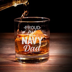 May include: A clear glass whisky tumbler with the words "Proud Navy Dad" etched on the side. The glass is filled with amber liquid and ice cubes. An American flag with an eagle is also etched on the glass.