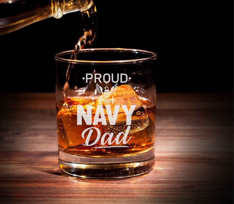 May include: A clear glass whisky tumbler with the words "Proud Navy Dad" etched on the side. The glass is filled with amber liquid and ice cubes. An American flag with an eagle is also etched on the glass.