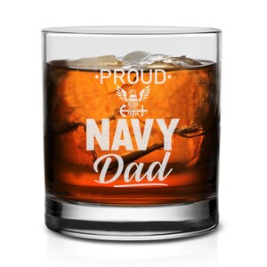 May include: A clear glass tumbler with a white design that reads "Proud Navy Dad" with an eagle and anchor graphic. The glass is filled with amber liquid and ice cubes.