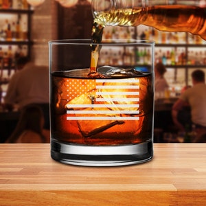 Puede incluir: Un vaso de rocas de vidrio transparente con un dise&ntilde;o de bandera estadounidense grabado en el lateral. El vaso est&aacute; lleno de l&iacute;quido &aacute;mbar y cubitos de hielo.