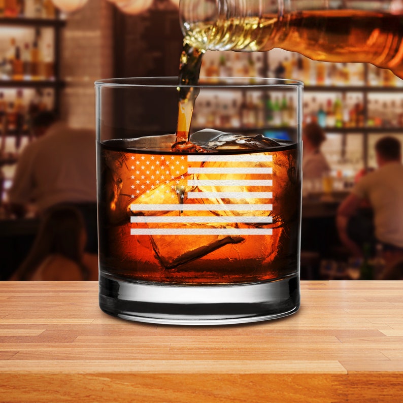 Puede incluir: Un vaso de rocas de vidrio transparente con un dise&ntilde;o de bandera estadounidense grabado en el lateral. El vaso est&aacute; lleno de l&iacute;quido &aacute;mbar y cubitos de hielo.