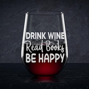 May include: A clear stemless wine glass with the text "Drink Wine Read Books Be Happy" printed on the side. The glass is filled with red wine.