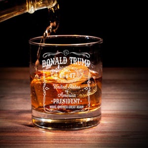 Puede incluir: Un vaso de whisky de vidrio transparente con el texto "DONALD TRUMP No. 45&47 United States of America &bull;PRESIDENT. MAKE AMERICA GREAT AGAIN" grabado en el lateral. El vaso est&aacute; lleno de l&iacute;quido &aacute;mbar y cubitos de hielo.