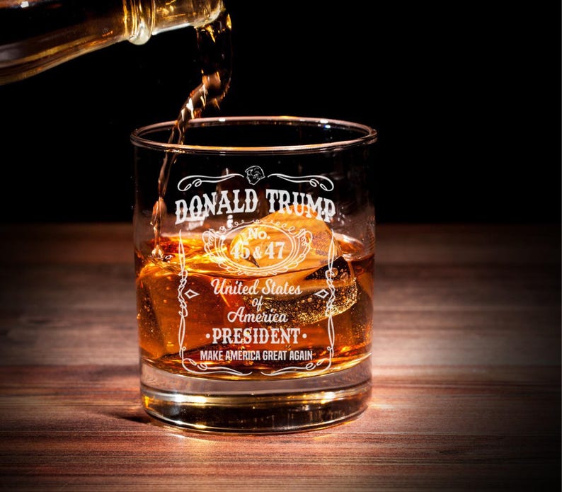 Puede incluir: Un vaso de whisky de vidrio transparente con el texto "DONALD TRUMP No. 45&47 United States of America &bull;PRESIDENT. MAKE AMERICA GREAT AGAIN" grabado en el lateral. El vaso est&aacute; lleno de l&iacute;quido &aacute;mbar y cubitos de hielo.
