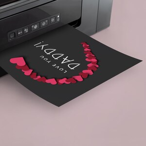 May include: A black card with the text "LOVE YOU DADDY" printed in white with a row of red paper hearts forming a semi-circle around the text. The card is being printed from a black printer.