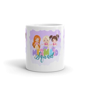 Puede incluir: Taza de cer&aacute;mica blanca con fondo morado y tres sirenas de dibujos animados. Las sirenas llevan colas de colores y tienen el pelo largo. El texto "Mermaid Squad" est&aacute; escrito en una fuente turquesa.