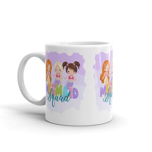 Puede incluir: Taza de cer&aacute;mica blanca con un dise&ntilde;o morado y blanco con tres sirenas de dibujos animados con el texto "Mermaid Squad".