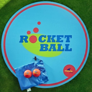 May include: A circular light blue game mat with a red border, featuring the text ROCKET BALL in blue letters over a lime green oval. Two orange balls sit on a blue drawstring bag, which also displays the Seaturtle Sports logo in red and yellow.