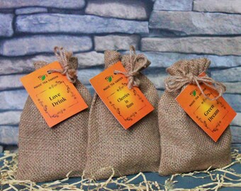 May include: Three small hessian bags with orange labels. The labels have the text "MAGIC TEA GARDEN" and the names of different tea blends: "Love Drink", "Cherries in Rum", and "Green Dream".