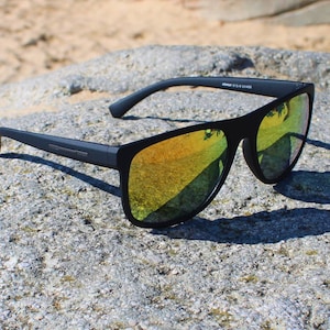 Könnte beinhalten: Schwarze Sonnenbrille mit rechteckigem Rahmen und gelb getönten, reflektierenden Gläsern. Die Sonnenbrille liegt auf einem grauen Felsen, mit sandigem Hintergrund. Die Sonnenbrille hat ein schlankes, modernes Design.