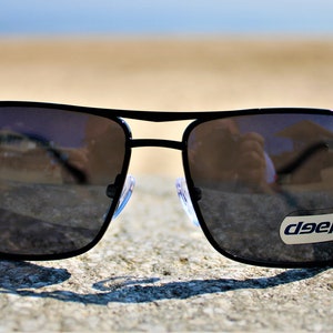 K&ouml;nnte beinhalten: Schwarze quadratische Sonnenbrille mit reflektierendem Glas. Die Sonnenbrille hat einen schwarzen Metallrahmen und ein kleines "deep"-Etikett auf der rechten Linse.