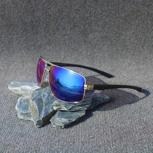 Könnte beinhalten: Eine Sonnenbrille mit goldfarbenem Rahmen und blauen und violetten verspiegelten Gläsern. Die Sonnenbrille liegt auf einem Stapel grauer Steine.