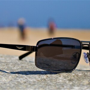May include: Black sunglasses with a dark gray lens and a silver frame. The sunglasses are sitting on a gray, textured surface. The sunglasses have a small logo on the side that says "Class".