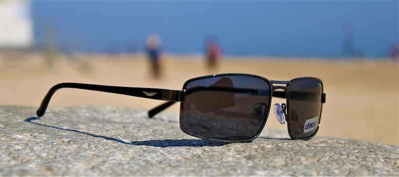 May include: Black sunglasses with a dark gray lens and a silver frame. The sunglasses are sitting on a gray, textured surface. The sunglasses have a small logo on the side that says "Class".