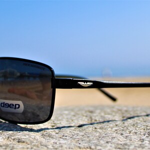 K&ouml;nnte beinhalten: Schwarze rechteckige Sonnenbrille mit einem silbernen Logo an der Seite. Die Sonnenbrille liegt auf einem grauen Stein mit einem Sandstrand und blauem Himmel im Hintergrund.