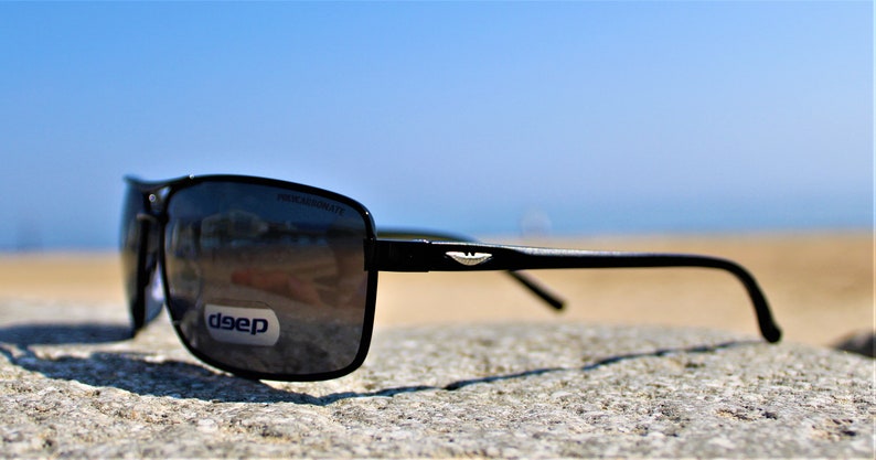 K&ouml;nnte beinhalten: Schwarze rechteckige Sonnenbrille mit einem silbernen Logo an der Seite. Die Sonnenbrille liegt auf einem grauen Stein mit einem Sandstrand und blauem Himmel im Hintergrund.
