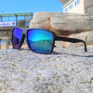 Könnte beinhalten: Schwarze Sonnenbrille mit blauen verspiegelten Gläsern. Die Gläser reflektieren eine Strandszene mit einem Pier im Hintergrund. Die Sonnenbrille ist aus Polycarbonat.