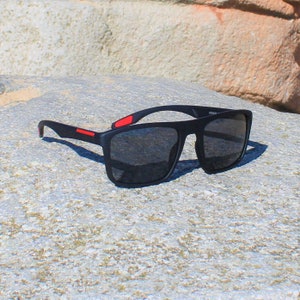 Könnte beinhalten: Schwarze quadratische Sonnenbrille mit roten Akzenten an den Bügeln. Die Sonnenbrille liegt auf einer grauen, strukturierten Oberfläche.