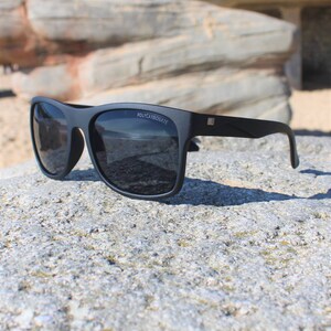 Könnte beinhalten: Schwarze rechteckige Sonnenbrille mit Polycarbonat-Gläsern. Die Sonnenbrille liegt auf einem grauen Stein.