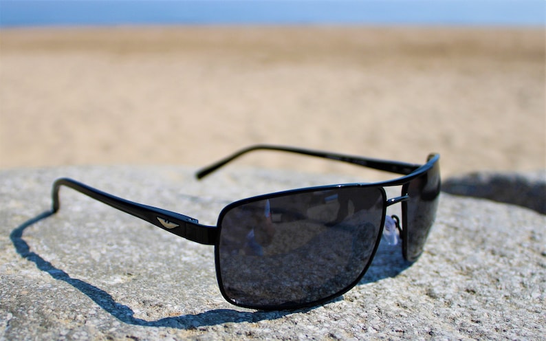 K&ouml;nnte beinhalten: Schwarze Sonnenbrille mit rechteckigem Gestell und dunklen Gl&auml;sern. Die Sonnenbrille liegt auf einem grauen Stein.