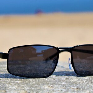 May include: Black rectangular sunglasses with a dark gray lens. The sunglasses are sitting on a gray rock.