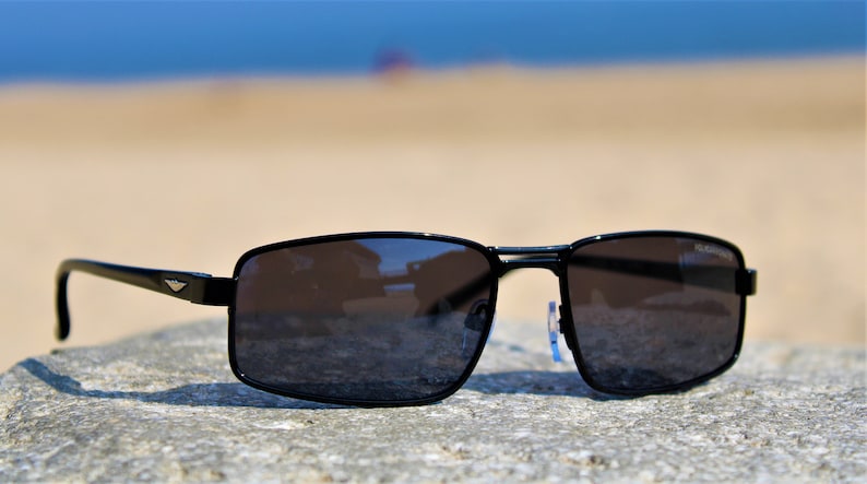 May include: Black rectangular sunglasses with a dark gray lens. The sunglasses are sitting on a gray rock.