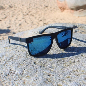Könnte beinhalten: Schwarze Sonnenbrille mit blauen Gläsern und einem schwarzen Rahmen. Die Sonnenbrille hat einen dezenten grün-roten Streifen an der Seite des Rahmens. Die Sonnenbrille liegt auf einem grauen Stein.