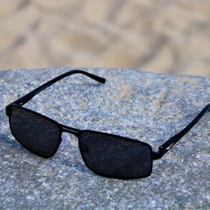 May include: Black rectangular sunglasses with dark lenses, resting on a gray textured surface.
