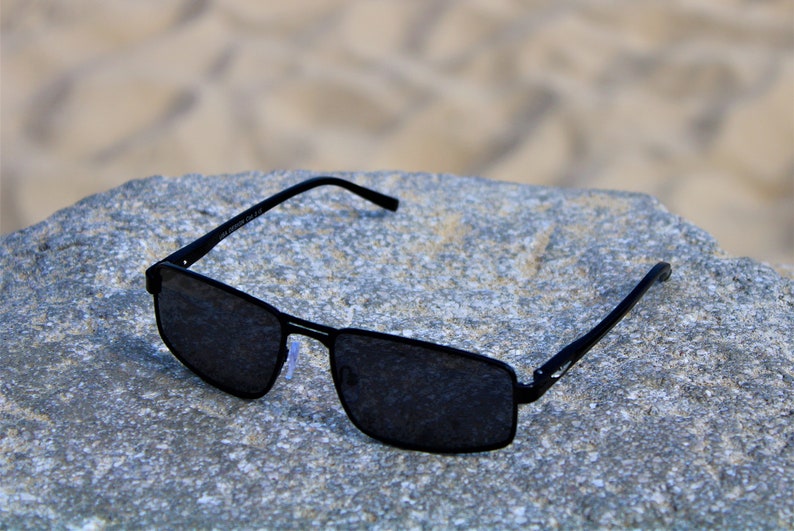 May include: Black rectangular sunglasses with dark lenses, resting on a gray textured surface.