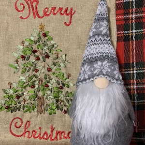 Peut inclure: Un gnome gris avec une barbe blanche et un chapeau à motifs gris et blancs. Le gnome est debout devant un sac en toile de jute avec les mots "Joyeux Noël" brodés dessus.