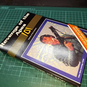 Könnte beinhalten: Eine schwarze und gelbe Schachtel mit dem Text "Revenge of the Apes" und "Games of the Century" auf der Vorderseite. Die Schachtel zeigt einen Mann, der eine Waffe in einer Dschungelkulisse hält. Der Text "Video Game Cartridge 11036" befindet sich an der Seite der Schachtel. Der Text "For Atari and Sears Video Game Systems" befindet sich auf der Rückseite der Schachtel.