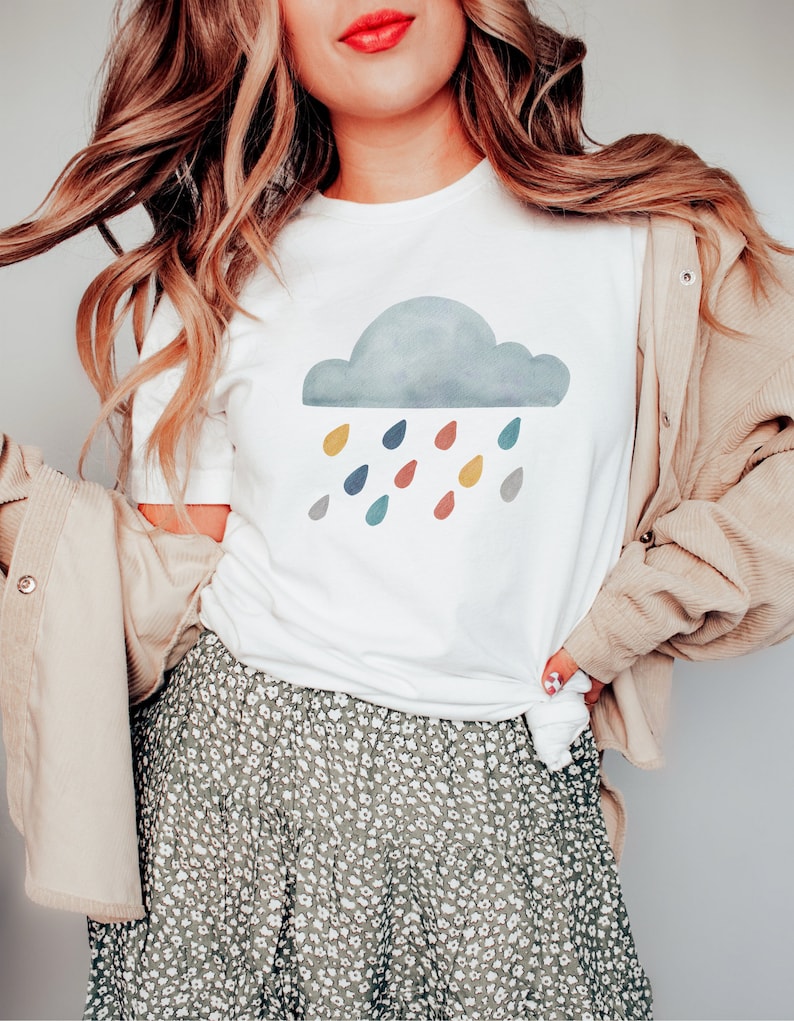 May include: A white t-shirt with a gray cloud and colorful raindrops graphic. The shirt is being worn by a person with long blonde hair and a floral skirt.