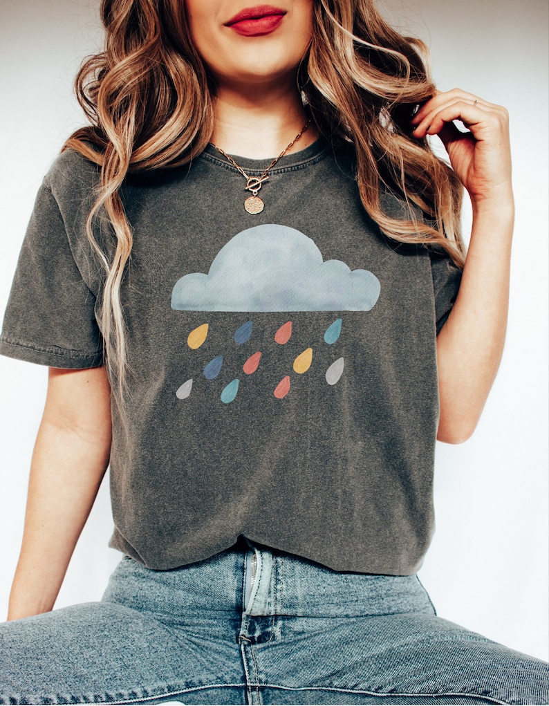May include: A dark gray t-shirt with a watercolor cloud and colorful raindrops graphic. The shirt has a relaxed fit and short sleeves.