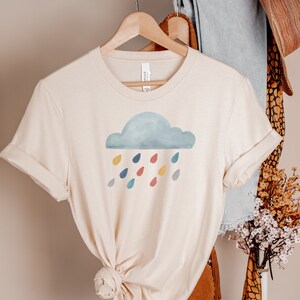 May include: A beige t-shirt with a watercolor cloud and colorful raindrops graphic. The shirt is on a hanger.