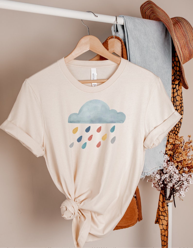 May include: A beige t-shirt with a watercolor cloud and colorful raindrops graphic. The shirt is on a hanger.