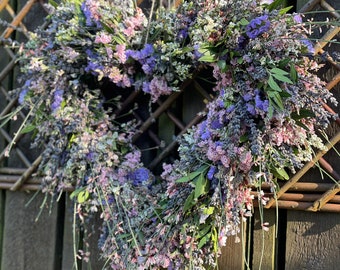 May include: A heart-shaped wreath made of dried flowers in shades of purple, pink, and green. The wreath is hanging on a wooden fence.