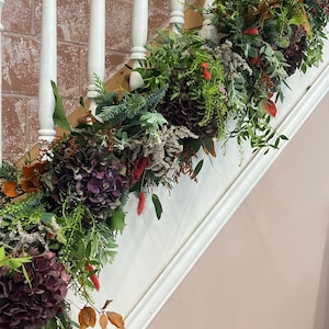 May include: A white wooden staircase banister decorated with a lush garland of greenery, burgundy hydrangeas, red berries, and brown leaves.