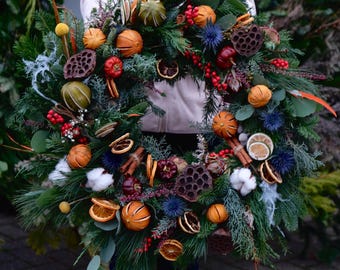 May include: A festive wreath featuring evergreen branches, dried oranges, cinnamon sticks, and red berries. The wreath is adorned with blue thistle flowers and various dried seed pods, creating a natural and seasonal decoration.