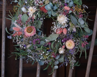 May include: A dried flower wreath with pink, white, and purple flowers, green leaves, and brown twigs. The wreath is hanging on a wooden fence.