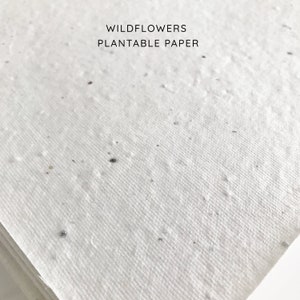以下が含まれることがあります： 黒い斑点のある白い植栽可能な紙のクローズアップ。紙には「WILDFLOWERS PLANTABLE PAPER」というテキストが印刷されています。