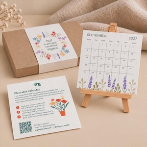 Pode incluir: Um conjunto de calendário plantável 2027 com uma caixa de cartão castanha com uma faixa floral, uma página de calendário quadrada para setembro num pequeno cavalete de madeira e um cartão de instruções com um código QR e flores.