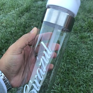 May include: A clear plastic water bottle with a white lid and a silver band around the middle. The bottle has a white straw and the word "Snazzy" written on the side in white cursive.
